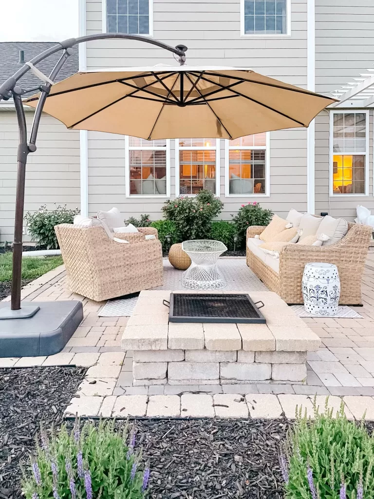 backyard scene with paver patio featuring firefit. stone is unilock cobblestone, brussels and hollandstone, patio furniture is better homes and garden 5 piece patio set from walmart. side tables are mixed with blue and white garden stool and vintage pieces. A cantilevel umbrella overarches the scene.